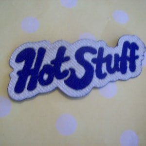 May include: An embroidered fabric patch featuring the text Hot Stuff in bold blue cursive lettering on a light gray background. The patch has a die-cut shape that follows the contour of the letters, creating a distinct and readable design.