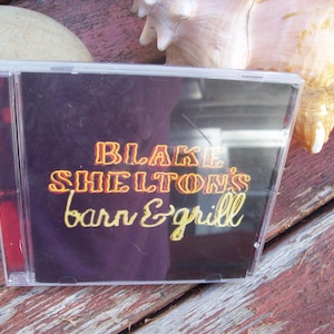 May include: A clear plastic CD case with a black background and the text "Blake Shelton's Barn & Grill" in yellow and orange letters.