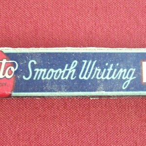 May include: A vintage Scripto pencil lead box. The box is rectangular with a blue background and the words "Scripto Smooth Writing LEADS" in white and red. The box is on a red surface.