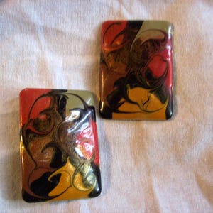 May include: Two rectangular, glossy earrings with abstract designs. The earrings feature a mix of colors, including red, black, gold, brown, and olive green. The earrings are set against a white background.