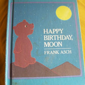 May include: A children's book titled "Happy Birthday, Moon" by Frank Asch. The cover features a brown bear silhouette on a brown hill, a yellow moon, and a blue starry sky background. The book has a blue border.