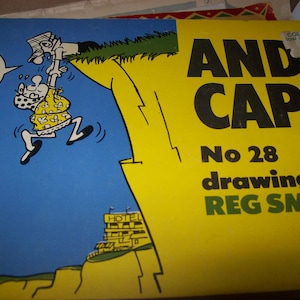 May include: A vintage "Andy Capp" comic book cover. The cover is yellow and blue, featuring a cartoon illustration of a character hanging from a cliff. Text includes "Andy Capp", "No 28", and "drawings by Reg Smythe".