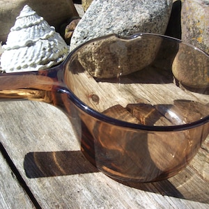 May include: A transparent amber-colored glass saucepan featuring a round bowl, a single integrated handle extending from the side, and a small pouring spout located on the rim. The glass is smooth and uniform in its brownish tint throughout the vessel.