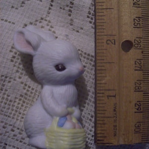 May include: A white ceramic bunny figurine holding a yellow basket with pastel-colored eggs. The bunny has pink inner ears and dark eyes. A wooden ruler with inch markings is next to the bunny. The ruler is approximately 3 inches long.