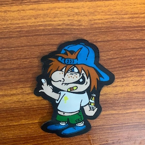 May include: A cartoon character patch featuring a figure with brown hair wearing a blue baseball cap, a white t-shirt with a yellow graphic, green shorts with the text VKJ, and blue shoes. The character holds a bottle and makes a hand gesture.