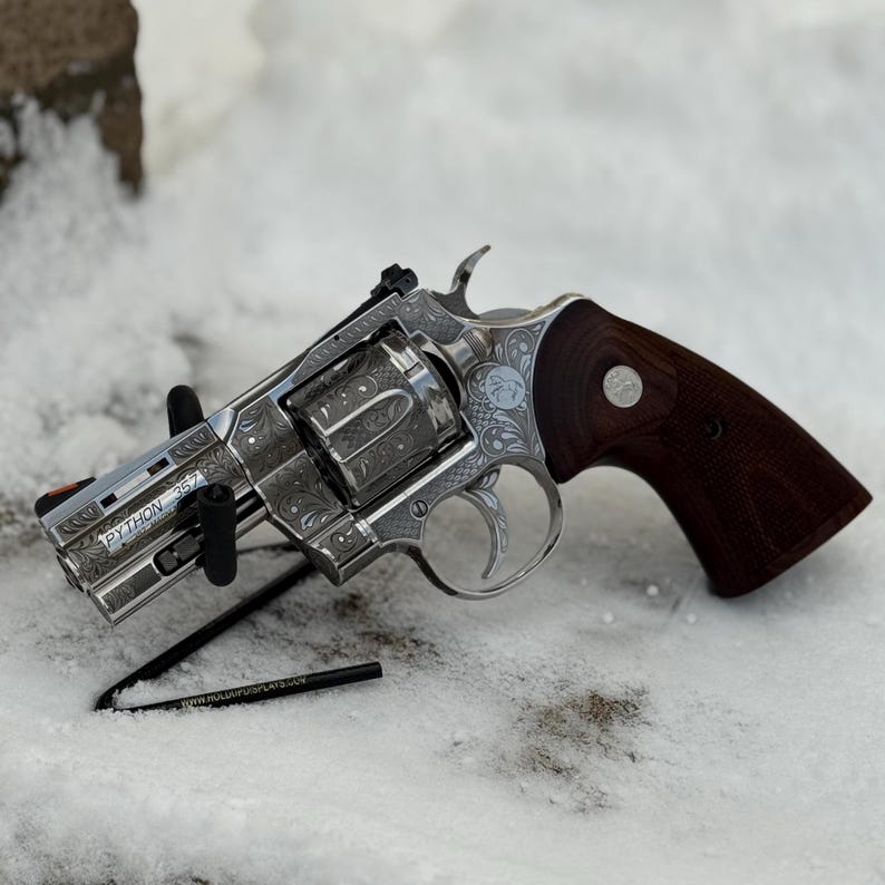 May include: A silver-toned revolver with intricate floral scrollwork engravings on the frame and cylinder. The firearm features a dark brown wooden grip with a circular emblem, a bright orange front sight, and the text PYTHON 357 on the barrel.