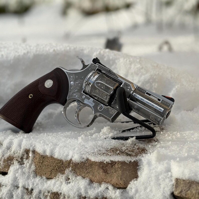 May include: A silver-toned revolver featuring intricate floral scrollwork engravings across the frame and cylinder. The firearm has a dark brown wooden grip with a circular emblem and a black front sight. Text on the barrel reads Colt's MFG. Hartford, CT. USA.
