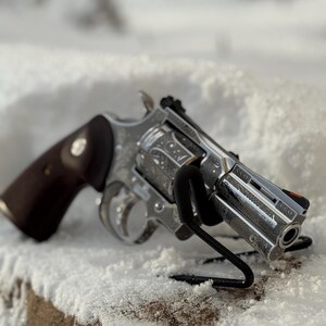 May include: A silver-colored revolver featuring intricate scrollwork engravings across the frame and cylinder. The firearm has a dark brown wooden grip with a circular emblem and a black front sight with an orange dot. Text on the barrel reads Colt's Mfg. Hartford, CT USA.