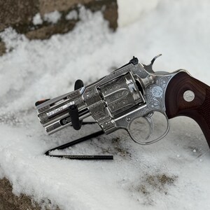 May include: A silver-toned revolver with intricate floral scrollwork engravings across the frame and cylinder. The barrel is marked with the text PYTHON 357. It features a dark brown wooden grip with a circular emblem and an orange front sight.