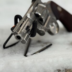 May include: A silver-toned revolver featuring intricate floral scrollwork engravings along the barrel and frame. The word PYTHON is clearly etched on the side of the barrel. It includes a bright orange front sight and dark wooden grips.