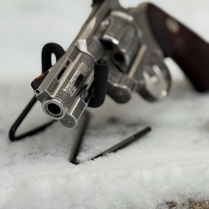 May include: A silver-toned revolver featuring intricate floral scrollwork engravings along the barrel and frame. The word PYTHON is clearly etched on the side of the barrel. It includes a bright orange front sight and dark wooden grips.