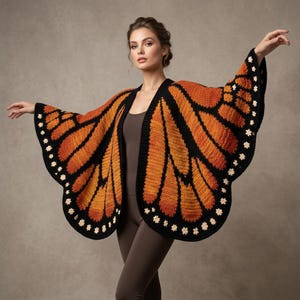 Könnte beinhalten: Ein gehäkelter Schal, der wie Monarchfalterflügel gestaltet ist, mit leuchtend orangefarbenen und schwarzen Garnmustern sowie kleinen weißen Blumenakzenten entlang der gewellten Ränder. Das Kleidungsstück wird über einem braunen Body und Leggings getragen.
