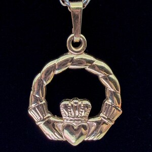May include: A gold-toned Claddagh pendant featuring a heart held by two hands, topped with a crown, and encircled by a twisted rope-like frame. The pendant is suspended from a matching gold-toned metal chain link necklace against a dark background.
