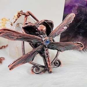 Può includere: Una collana con un ciondolo a forma di libellula in rame, con una pietra preziosa blu al centro. La libellula è dettagliata con ali intricate e un corpo delicato.