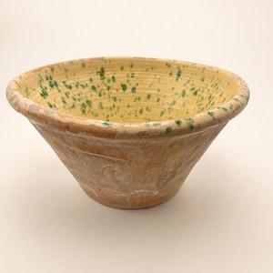 May include: A wide, conical earthenware bowl with a rustic, unglazed terracotta exterior. The interior features a pale yellow glaze with a speckled pattern of green paint splatters and a worn, chipped rim showing the natural clay underneath.
