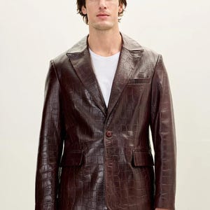 Peut inclure: Un blazer marron foncé avec un motif texturé en relief façon crocodile, un revers cranté, une fermeture à un bouton sur le devant et deux poches à rabat à la taille. Le vêtement est porté sur un t-shirt blanc uni et un pantalon sombre.