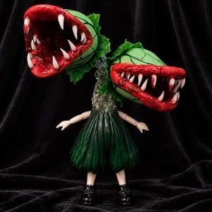 Puede incluir: Una figura de criatura vegetal de dos cabezas con cabezas bulbosas verdes veteadas, interiores rojos y dientes blancos afilados. La figura tiene un cuello texturizado y musgoso, una falda plisada verde oscuro, brazos articulados y zapatos negros.