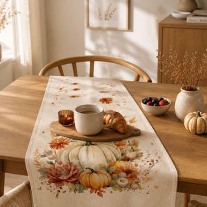 Könnte beinhalten: Ein cremefarbener Tischläufer aus Stoff mit einem Druckmotiv aus orangefarbenen und weißen Kürbissen, Herbstblättern und Blumen. Der Läufer liegt auf einem Holztisch mit einer Keramiktasse, einem Croissant, einer Kerze und Beeren.