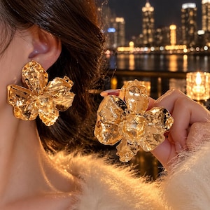 May include: A pair of large, gold-toned earrings shaped like blooming flowers with a textured, hammered metal finish. One earring is attached to an earlobe, while the other is held up to display the detailed, irregular petal design.