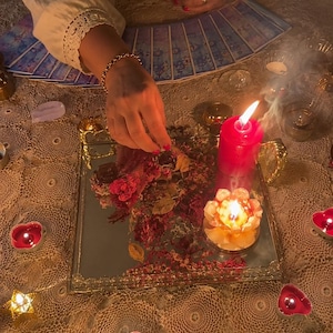 May include: A square mirrored tray holds dried flower petals, a tall red pillar candle, and a lotus-shaped candle. The tray is surrounded by small heart-shaped and star-shaped tea light candles on a lace tablecloth with a fan of tarot cards.