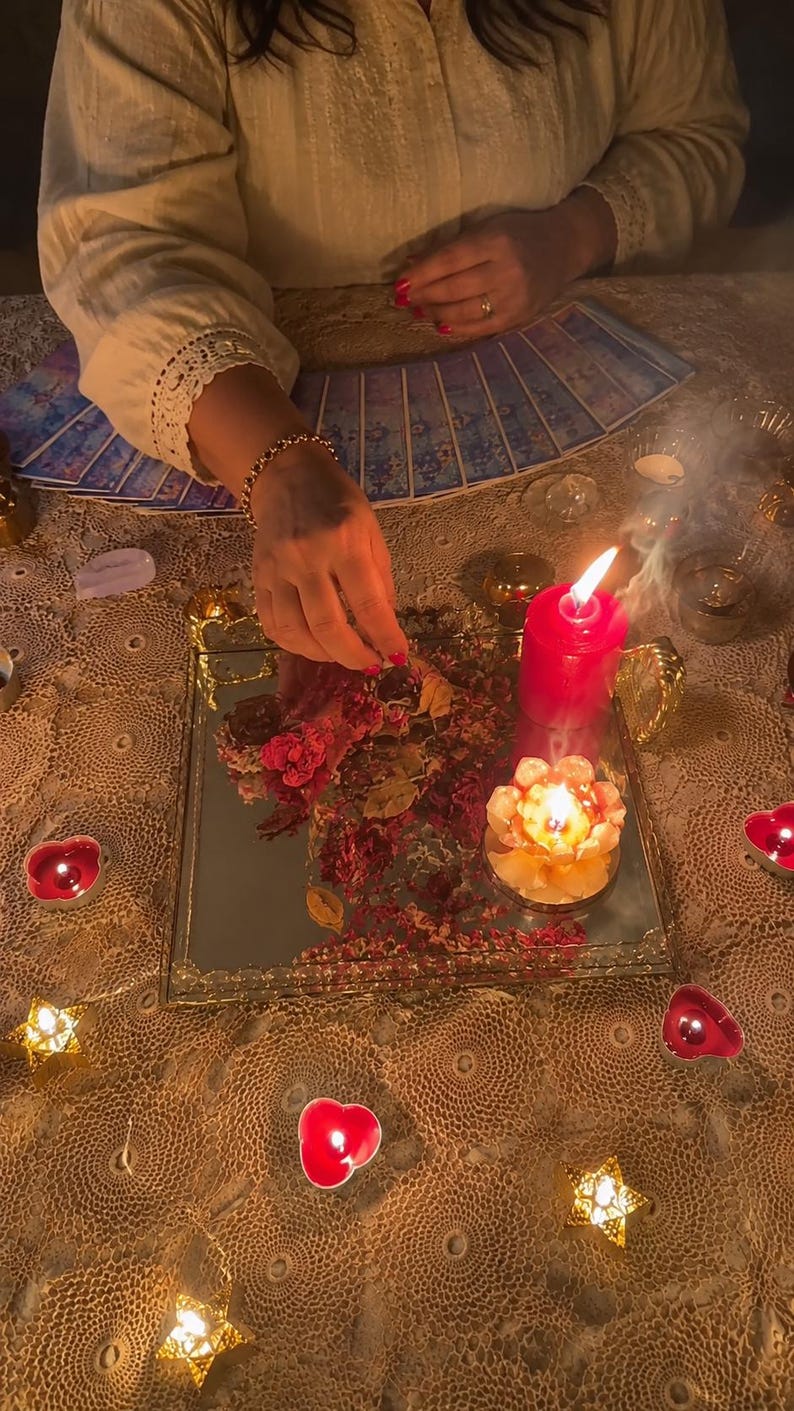 May include: A square mirrored tray holds dried flower petals, a tall red pillar candle, and a lotus-shaped candle. The tray is surrounded by small heart-shaped and star-shaped tea light candles on a lace tablecloth with a fan of tarot cards.