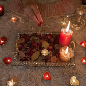 May include: A rectangular silver tray with crystal-like edges holds dried red rose petals, bay leaves, and smooth stones. Two lit pillar candles, one red and one white, sit on the tray alongside a small tea light candle, surrounded by heart-shaped candles.