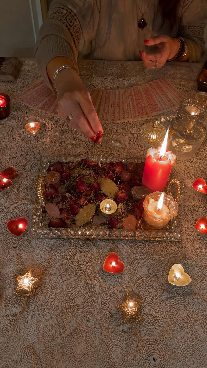 May include: A rectangular silver tray with crystal-like edges holds dried red rose petals, bay leaves, and smooth stones. Two lit pillar candles, one red and one white, sit on the tray alongside a small tea light candle, surrounded by heart-shaped candles.