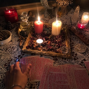 May include: A spread of tarot cards with pink patterned backs arranged on a white lace tablecloth. A gold-toned tray filled with dried flower petals, a red pillar candle, a white pillar candle, and a small tea light candle sits in the center.