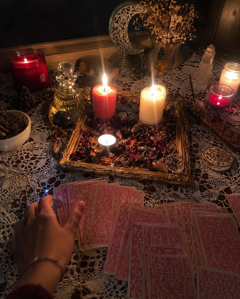 May include: A spread of tarot cards with pink patterned backs arranged on a white lace tablecloth. A gold-toned tray filled with dried flower petals, a red pillar candle, a white pillar candle, and a small tea light candle sits in the center.