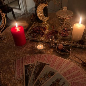 May include: A spread of tarot cards featuring the Queen of Cups and other figures, arranged on a lace tablecloth. The scene includes a red pillar candle, a white pillar candle, a gold crescent moon sculpture, and a glass tray with dried petals.