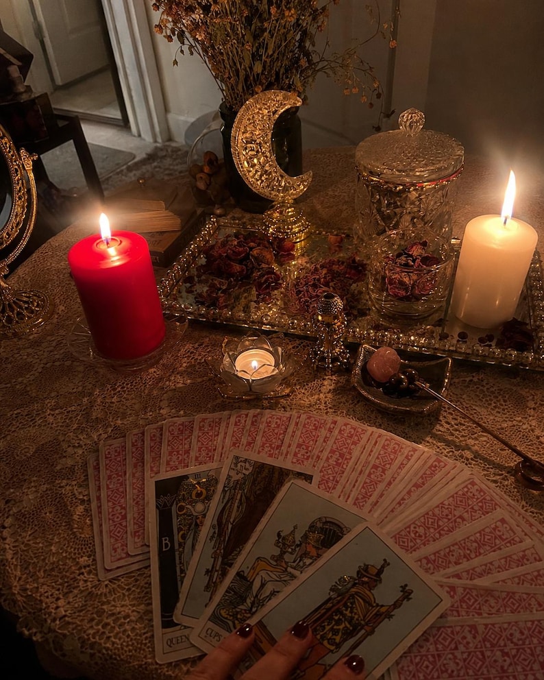 May include: A spread of tarot cards featuring the Queen of Cups and other figures, arranged on a lace tablecloth. The scene includes a red pillar candle, a white pillar candle, a gold crescent moon sculpture, and a glass tray with dried petals.