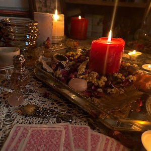 May include: A rectangular mirrored tray holds a large red pillar candle, dried flower petals, small yellow blossoms, and smooth pink stones. Nearby are a small glass perfume bottle, a silver spoon, and a row of pink patterned tarot cards.