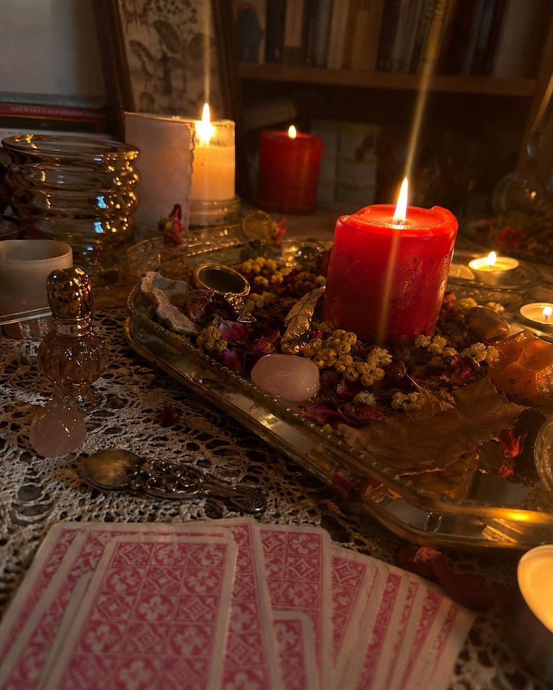 May include: A rectangular mirrored tray holds a large red pillar candle, dried flower petals, small yellow blossoms, and smooth pink stones. Nearby are a small glass perfume bottle, a silver spoon, and a row of pink patterned tarot cards.