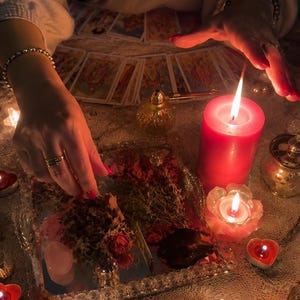 May include: A collection of tarot cards arranged in a fan shape, a large red pillar candle, a small lotus-shaped candle, and several heart-shaped tea light candles. A mirrored tray holds dried herbs, rose petals, and a small pink crystal stone.