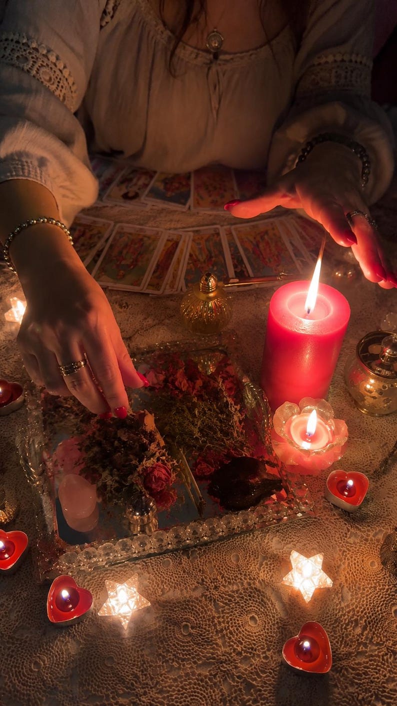 May include: A collection of tarot cards arranged in a fan shape, a large red pillar candle, a small lotus-shaped candle, and several heart-shaped tea light candles. A mirrored tray holds dried herbs, rose petals, and a small pink crystal stone.