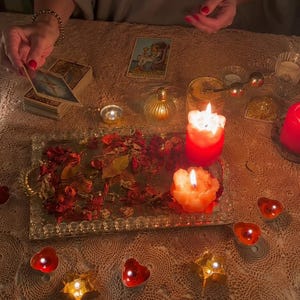 May include: A rectangular mirrored tray filled with dried red rose petals, positioned among several lit candles in red and orange hues. The scene includes a deck of tarot cards, a single Queen of Cups card, and small star-shaped candle holders.