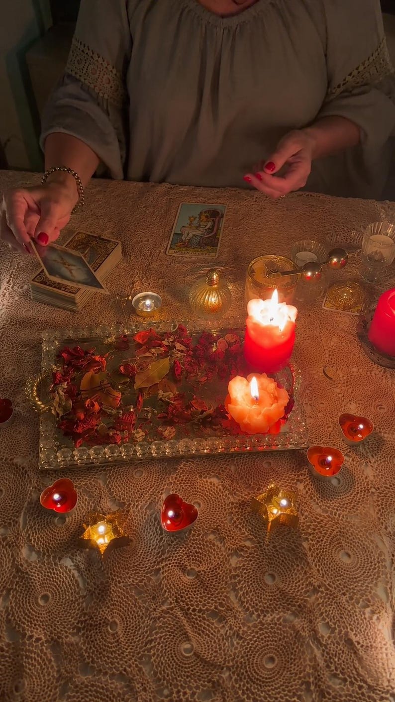May include: A rectangular mirrored tray filled with dried red rose petals, positioned among several lit candles in red and orange hues. The scene includes a deck of tarot cards, a single Queen of Cups card, and small star-shaped candle holders.