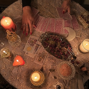 May include: A collection of tarot cards spread across a lace-covered table, accompanied by several lit candles in various holders, a silver tray filled with dried rose petals, a small bowl of pink salt crystals, and a white crescent moon ornament.