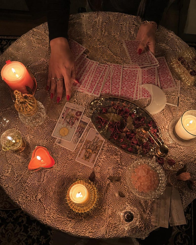 May include: A collection of tarot cards spread across a lace-covered table, accompanied by several lit candles in various holders, a silver tray filled with dried rose petals, a small bowl of pink salt crystals, and a white crescent moon ornament.