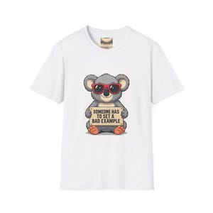 May include: A white short-sleeved t-shirt featuring a graphic of a grey koala wearing red sunglasses. The koala is holding a tan rectangular sign with the black text "SOMEONE HAS TO SET A BAD EXAMPLE" printed in a bold, sans-serif font.