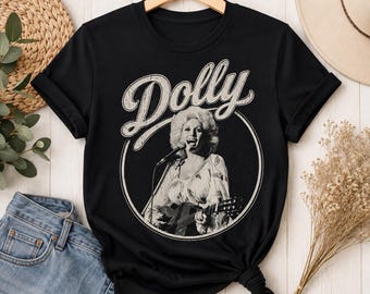 Può includere: Una t-shirt nera a maniche corte con una stampa grafica circolare di una persona che canta con una chitarra e un microfono. La parola Dolly è stampata in un grande carattere corsivo bianco invecchiato sopra l'immagine circolare.