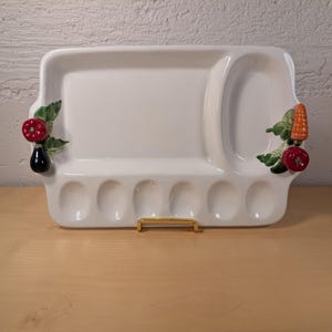 May include: A white ceramic serving tray featuring a large rectangular section, an oval section, and six small circular indentations. The tray is decorated with three-dimensional ceramic vegetables including a red pepper, eggplant, carrot, and tomato.