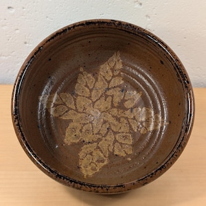 May include: A round, brown ceramic bowl with a speckled finish and a textured leaf pattern imprinted in the center. The interior of the bowl features concentric circular ridges, and the rim is accented with a dark, mottled brown glaze.