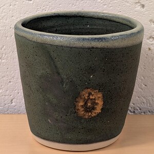 May include: A dark green ceramic cup with a speckled texture and a smooth, lighter green rim. The exterior features a single, circular, textured brown floral-like imprint on the side. The base of the cup is a contrasting light cream color.