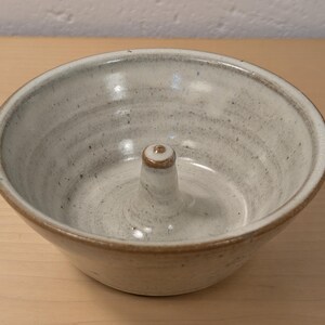 May include: A round ceramic bowl with a speckled off-white glaze and a brown rim. The interior features a central, tapered vertical post with a brown tip, creating a functional design for holding items like fruit or other small objects securely.