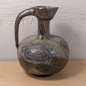 May include: A ceramic pitcher with a rounded body and a curved handle. The surface features a speckled beige and brown glaze with embossed floral patterns in blue and green. The neck is narrow, leading to a flared rim with a small pouring spout.