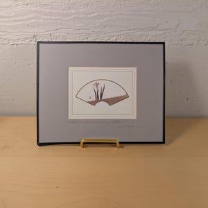 May include: A rectangular framed print featuring a central illustration of a fan shape containing a purple iris flower and green leaves. The print is set against a gray mat with a thin black border and includes handwritten text below the image.