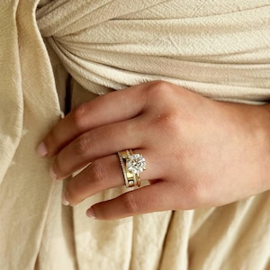 Puede incluir: Un conjunto de anillos de oro que presenta una gran piedra central redonda de talla brillante sujeta por garras, acompañada por una banda de oro más ancha con una fila de pequeños diamantes en pavé y una banda de oro más fina, con acabado pulido.