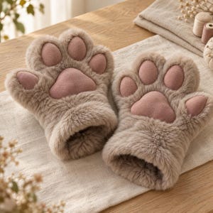 Op de afbeelding: Een paar vingerloze handschoenen van zacht, beige imitatiebont. Elke handschoen heeft een ontwerp van lichtroze, suède-achtig materiaal op de handpalm, met vier afgeronde teenkussentjes en één groter, centraal kussentje in de vorm van een poot.