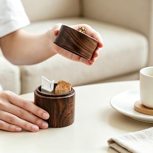 Puede incluir: Un recipiente cilíndrico de madera marrón oscuro con una tapa extraíble que tiene un pequeño pomo de metal dorado. El recipiente contiene una colección de palillos de hilo dental de plástico blanco y un grupo de palillos de madera clara.